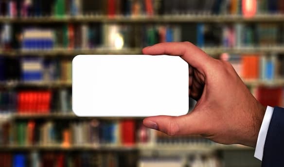 De fondo una estanteria con varios libros y la mano de una persona sosteniendo un teléfono móvil en primer plano